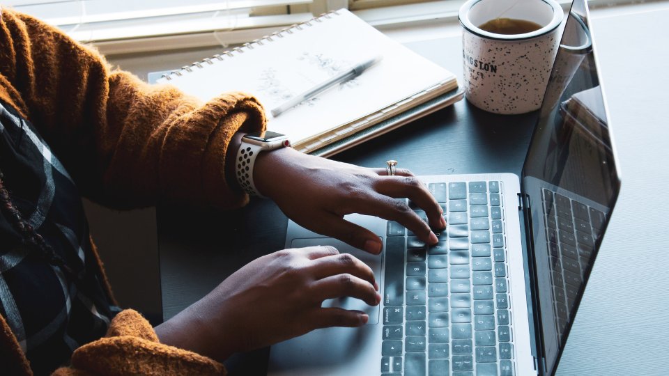 A person typing on a laptop in front of a cup of coffee.