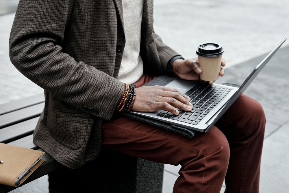 A person working on a laptop outdoors with coffee in their hand.