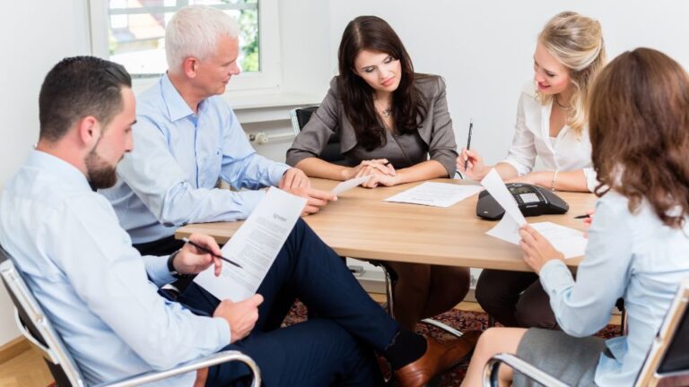 A group of colleagues engaged in a discussion while sitting around a table, looking at documents.