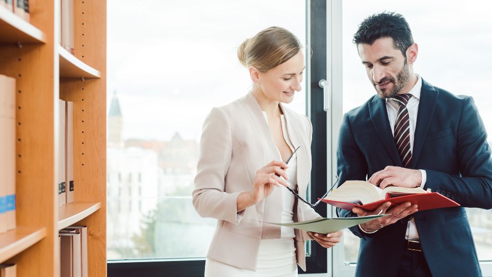 Two coworkers having a conversation, with a book and paperwork in their hands.