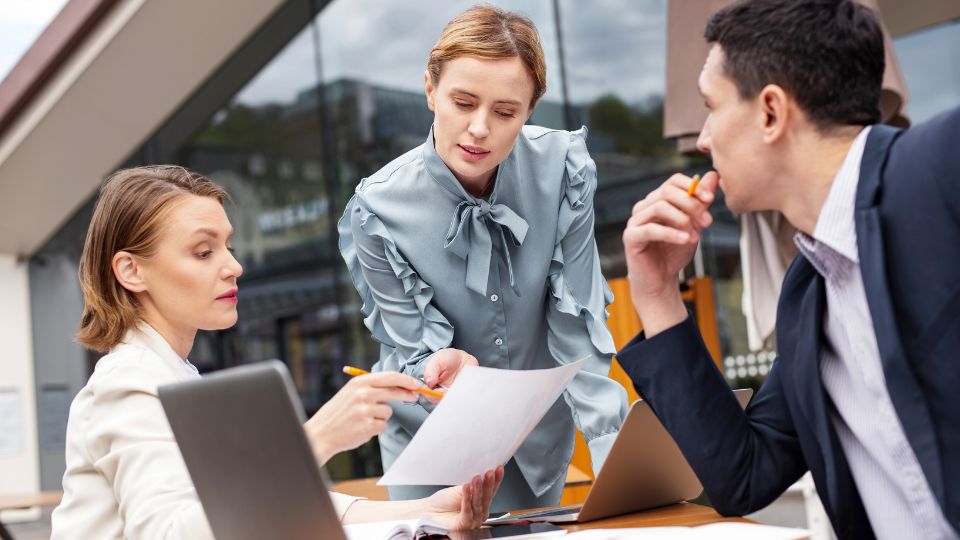 Three professionals are sitting at a table with laptops open and discussing a document.