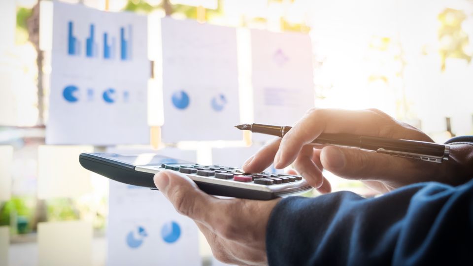 A person using a calculator with multiple printed financial charts and blue graphs on a wall in the background.