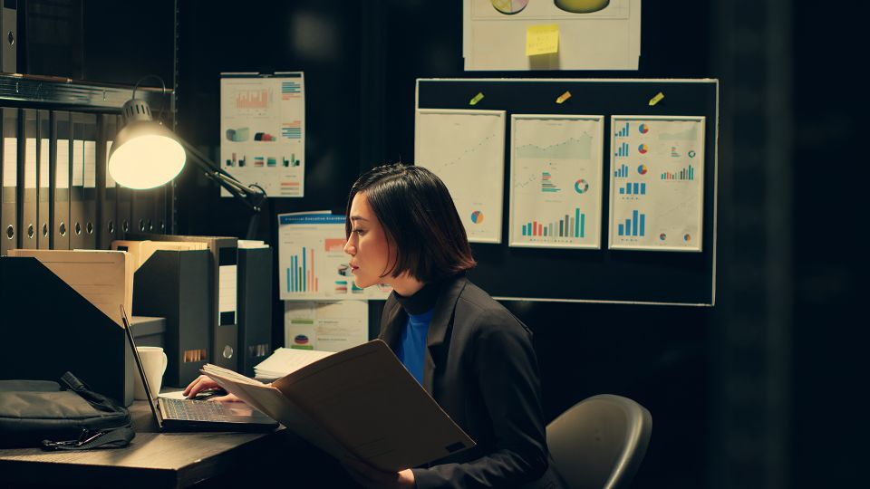 A professional reviewing data and reports on a laptop in a dimly lit office, surrounded by charts and documents