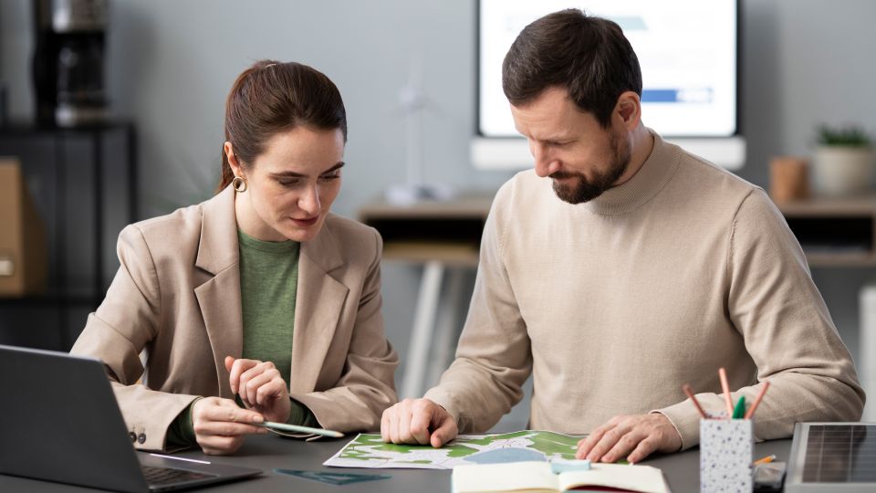 Two professionals review documents beside a laptop in an office setting.