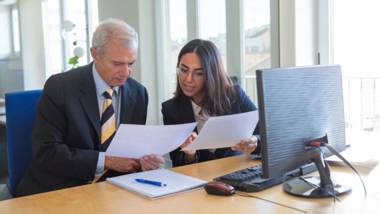 Two business professionals sit side-by-side at an office desk, reviewing documents in front of a computer.