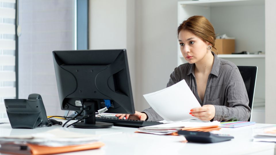 A business professional sits at an office desk, looking intently at a computer monitor while holding a piece of paper in her hand.