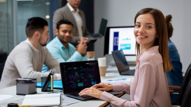 A woman working on a laptop displaying data charts while her colleagues discuss a project in the background.