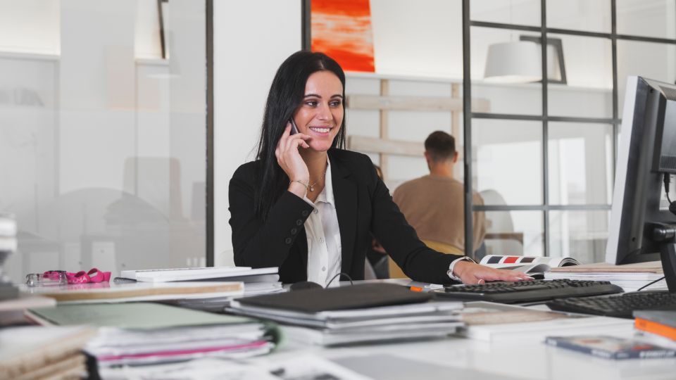 A smiling professional talks on a phone while sitting at an office desk.