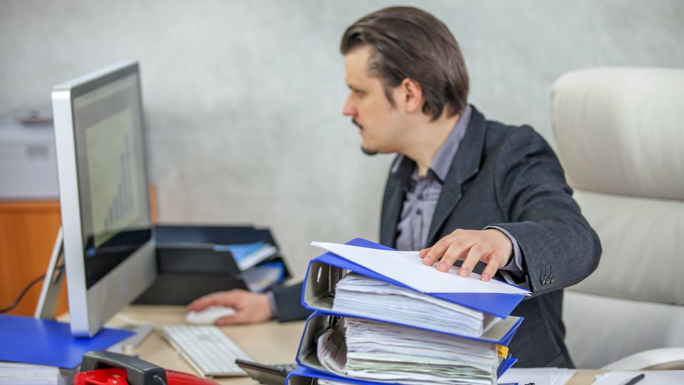 An individual reaches out to pick up a stack of documents in blue binders from a desk while looking at a computer monitor.