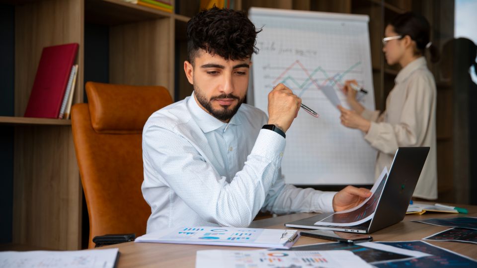 A focused man sits in a chair, looking at his laptop screen with several reports on the desk
