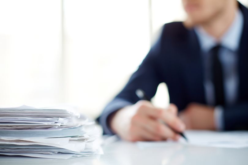 A close-up image of a stack of papers with a person in a suit in the background.
