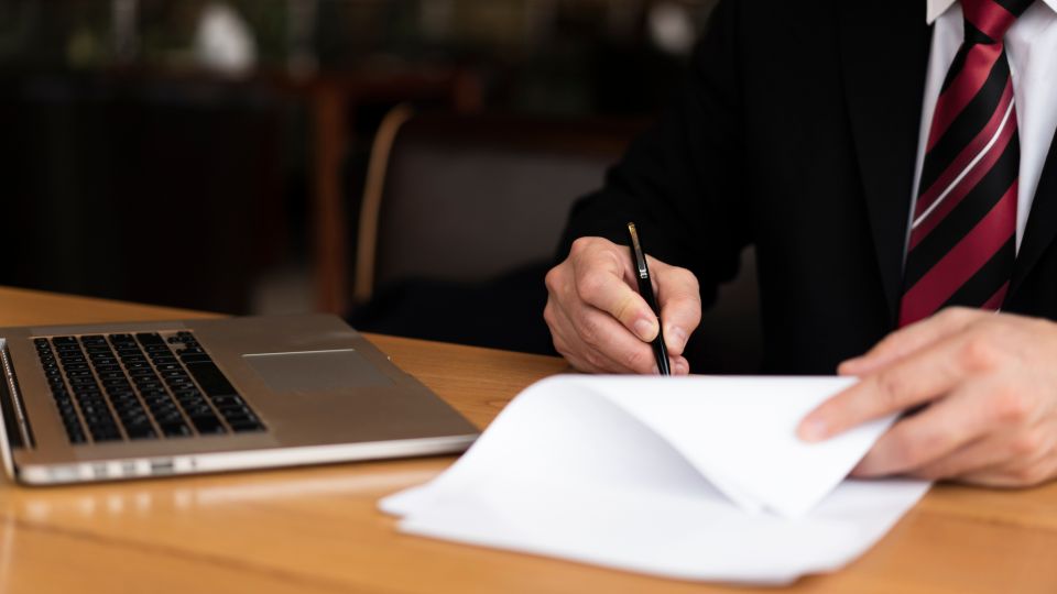 An individual sits at a wooden table with a laptop and signs a document with a black pen.