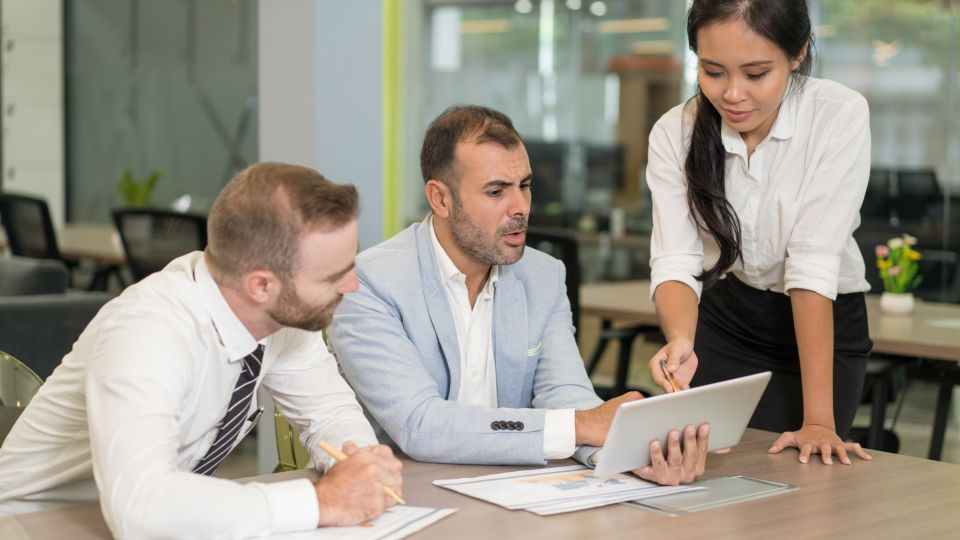 Three co-workers are analyzing data and documents on a tablet in an office.