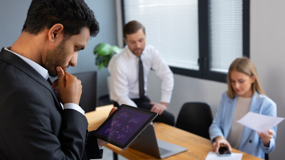 A man in a black suit looks down at a tablet displaying various graphs and charts.