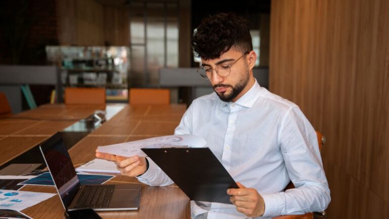 A professional sits at a large wooden table with a laptop, reviewing papers on a clipboard.