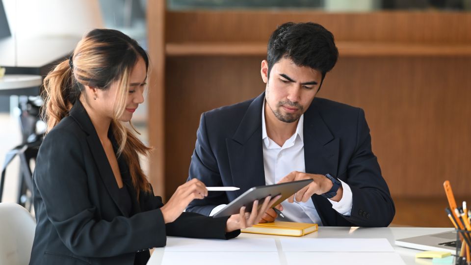 Two business professionals sit at a desk reviewing information on a laptop