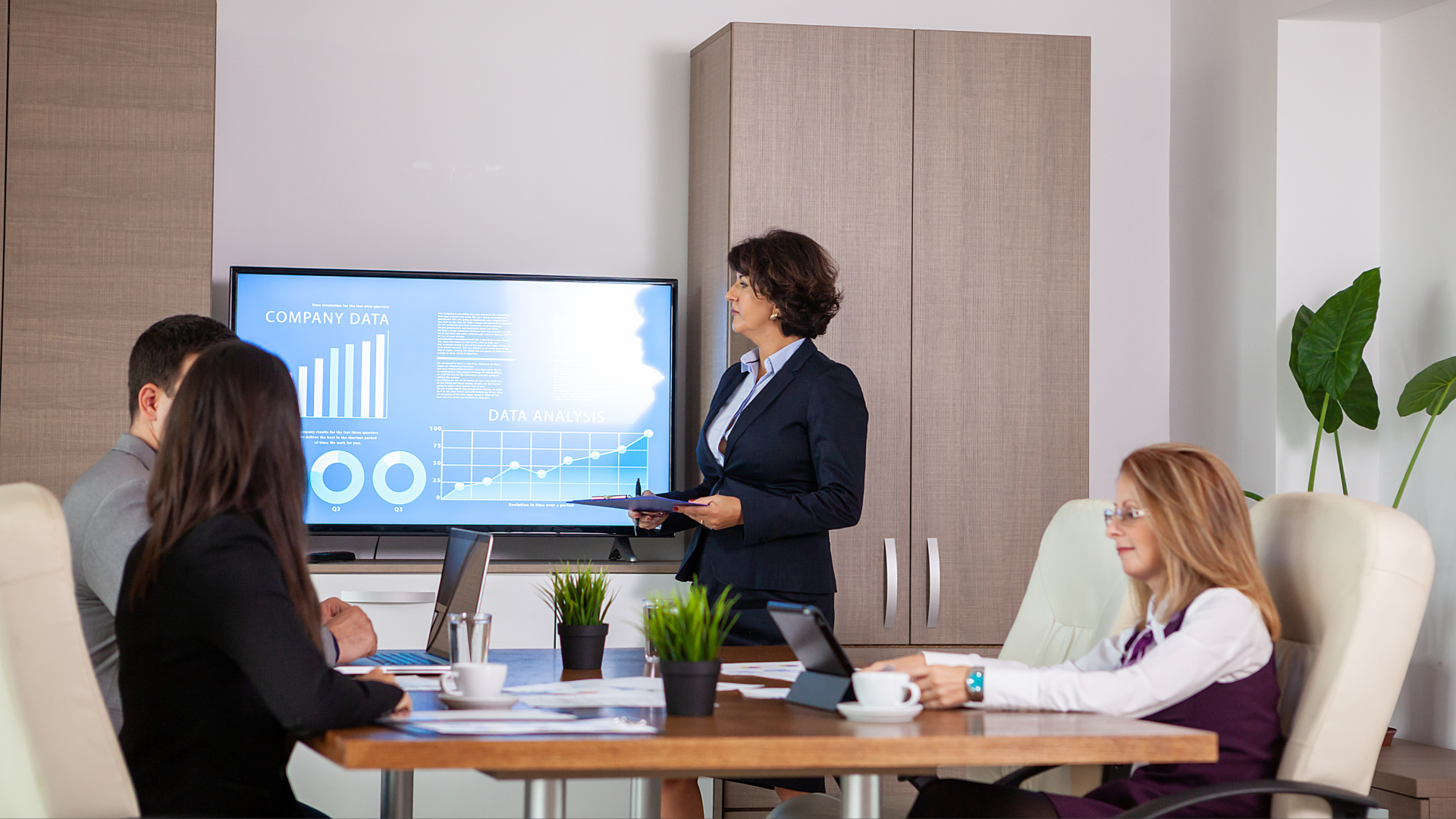 A woman stands by a large monitor displaying data charts while presenting to colleagues in a conference room.